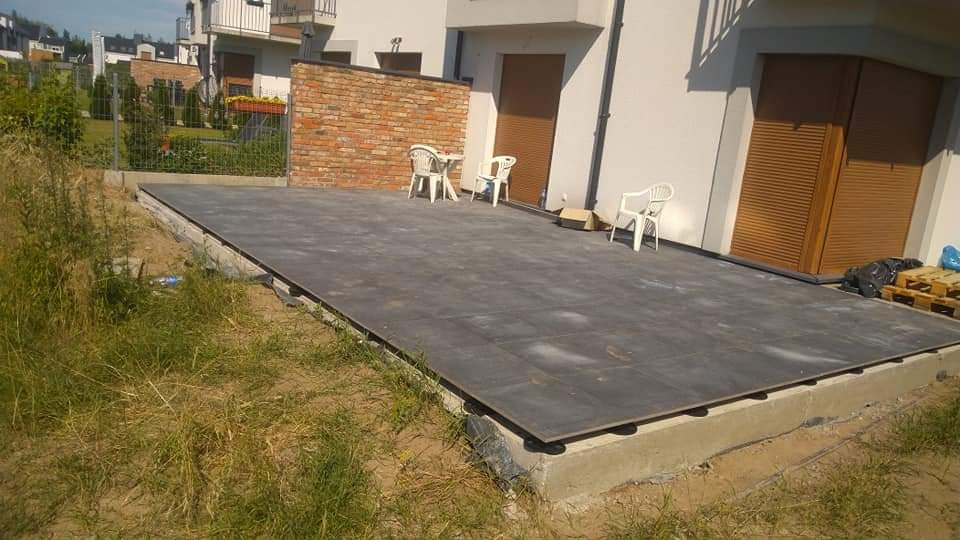 Nowoczesny taras z ciemnych płyt na betonowej podbudowie, widoczne podkładki dystansowe, w tle budynek mieszkalny z ceglanym murem i białymi krzesłami ogrodowymi.