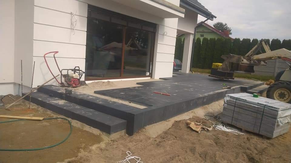 Częściowo ułożona, ciemnoszara kostka brukowa na tarasie domu jednorodzinnego, z widocznym sprzętem budowlanym i stertą kostki w opakowaniu, w tle jasna elewacja budynku z dużym oknem.