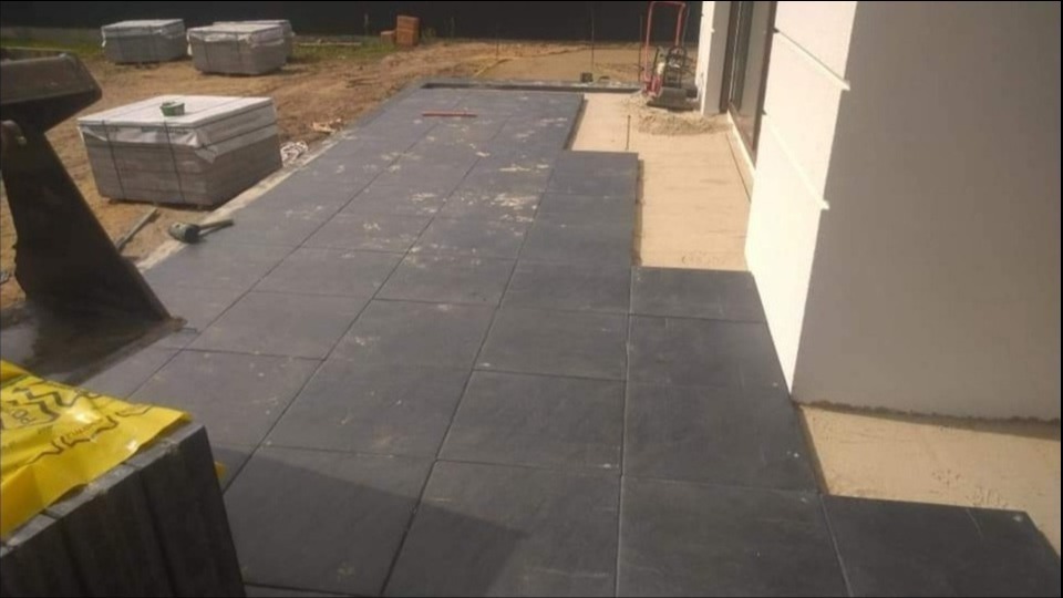 Częściowo ułożona, ciemnoszara nawierzchnia z kwadratowych płyt betonowych przylegająca do białej ściany budynku, z widocznymi przygotowanymi podłożem z piasku i narzędziami budowlanymi w tle.