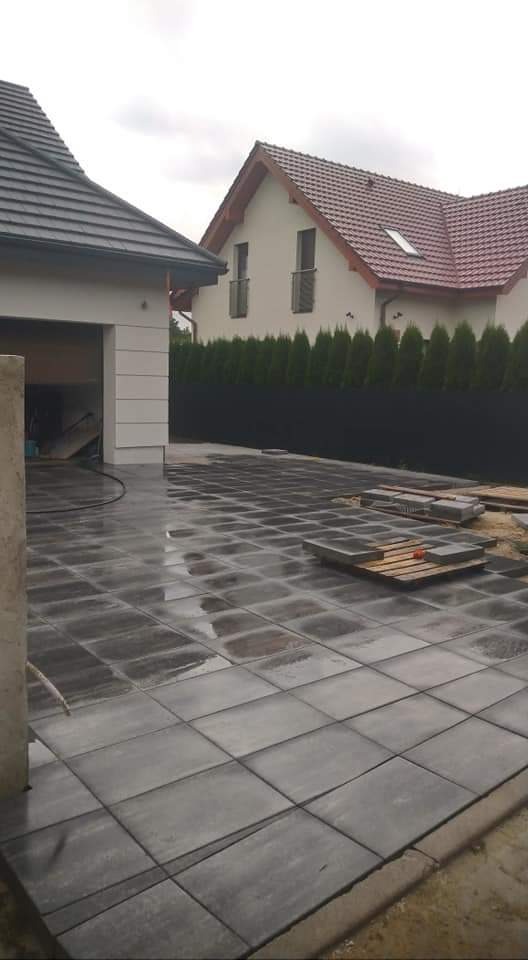 Świeżo ułożona, mokra, grafitowa kostka brukowa na podjeździe, z paletami kostki w tle, widok na dom z garażem i rząd tuj za czarnym ogrodzeniem.