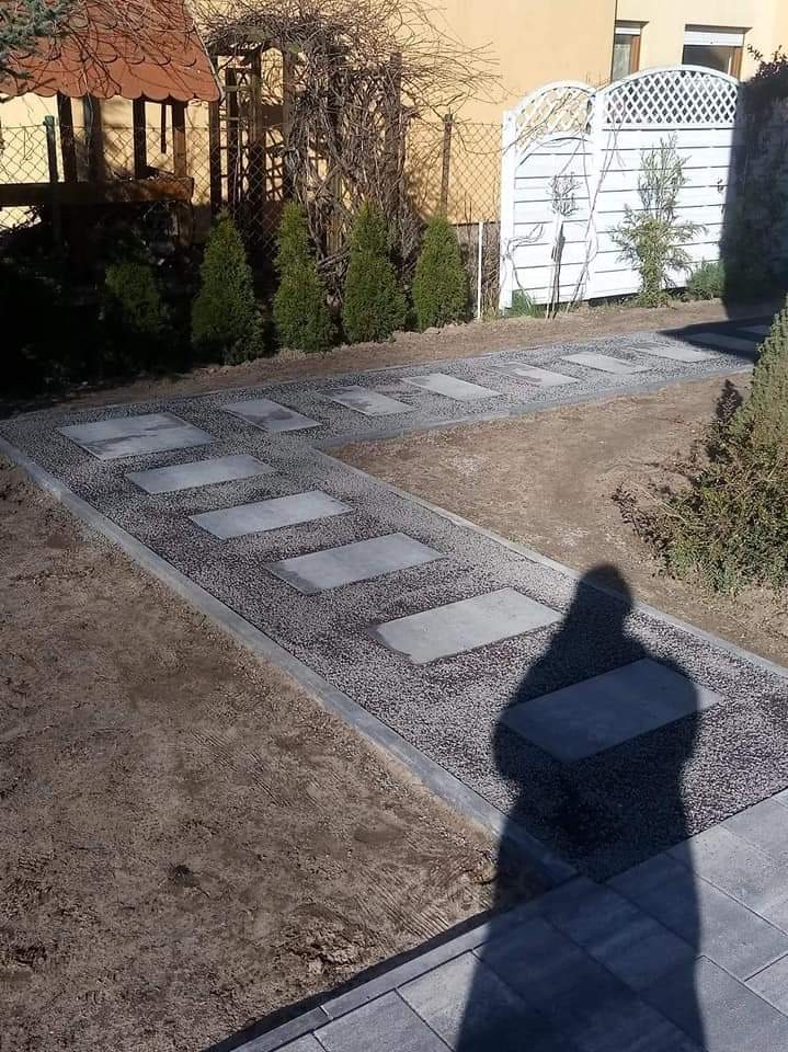 Ścieżka w ogrodzie z płyt betonowych ułożonych w szarym żwirze, obrzeża z kostki, w tle zielone tuje i biała ażurowa ścianka, cień osoby robiącej zdjęcie.