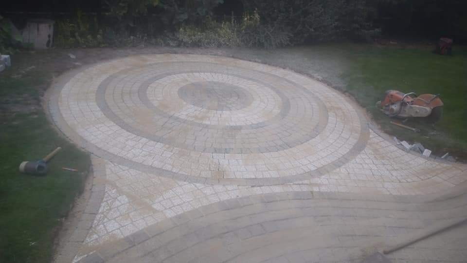 Układanie kostki granitowej w koncentryczne okręgi na trawniku, widoczny młotek brukarski i przecinarka do kamienia, etap prac wykończeniowych.