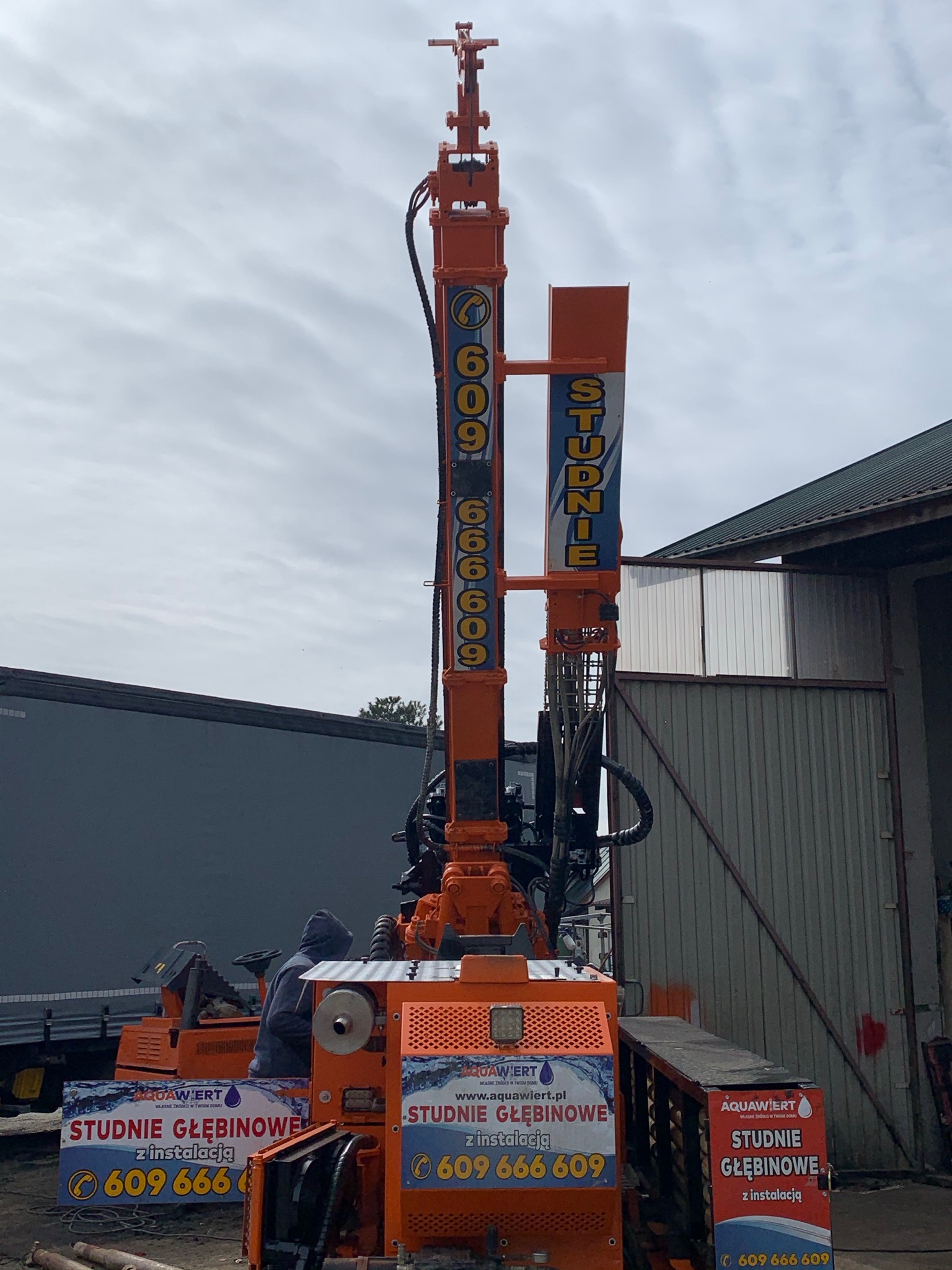 Pomarańczowa wiertnica do studni głębinowych z widocznym operatorem w szarej bluzie z kapturem, na tle szarej ściany budynku gospodarczego i pochmurnego nieba.