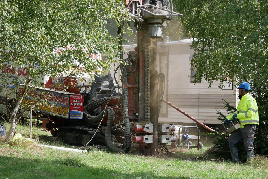 Wiercenie studni głębinowej w Międzybrodziu Bialskim przy użyciu ciężkiego sprzętu wiertniczego; w tle widoczny biały przyczep kempingowy, a pracownik w kasku i odblaskowej kurtce obsługuje...