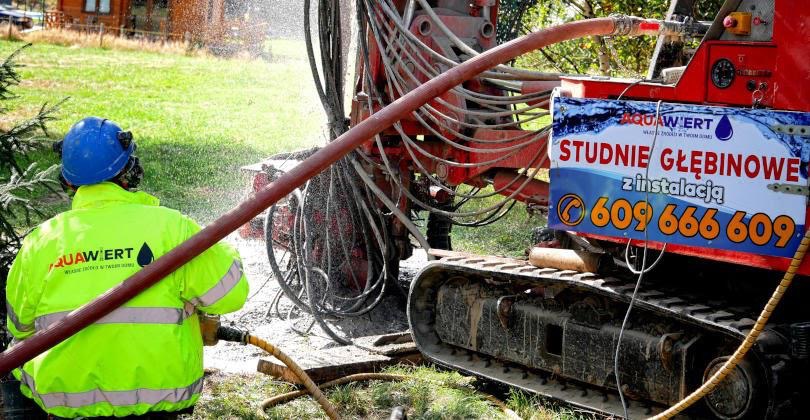 Pracownik w neonowej kurtce obsługuje wiertnicę gąsienicową podczas wiercenia studni; w tle woda rozpryskuje się na trawnik.