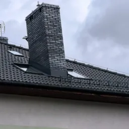 Nowy dach z czarnej dachówki ceramicznej, z oknami połaciowymi i kominem z ciemnej cegły klinkierowej, na tle pochmurnego nieba.