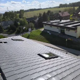 Nowe, szare pokrycie dachowe z widocznymi oknami dachowymi, w tle zabudowania i zielone pola pod błękitnym niebem z chmurami.