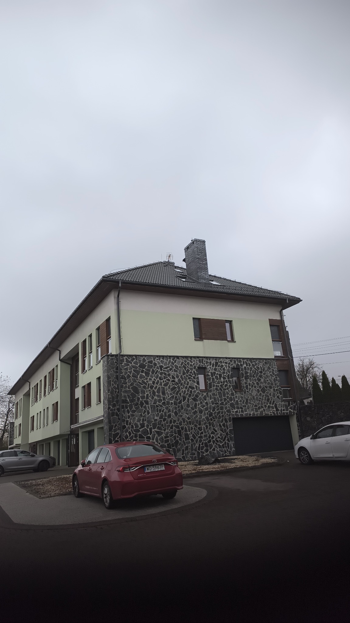 Dwukondygnacyjny budynek z szarym dachem, jasnymi ścianami i kamienną elewacją do połowy wysokości, widziany z perspektywy ulicy w pochmurny dzień.