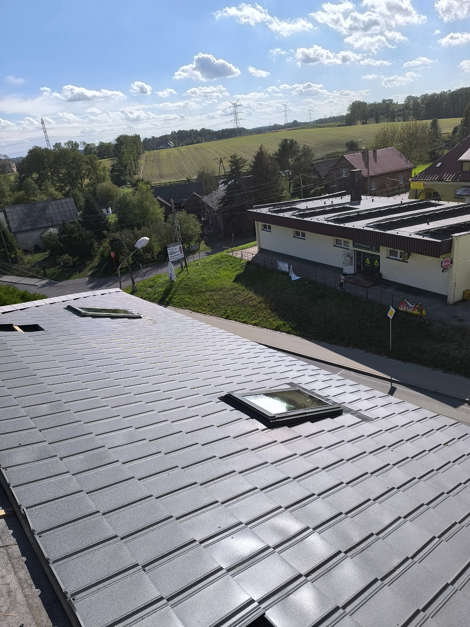 Nowe, szare pokrycie dachowe z widocznymi oknami dachowymi, w tle zabudowania i zielone pola pod błękitnym niebem z chmurami.