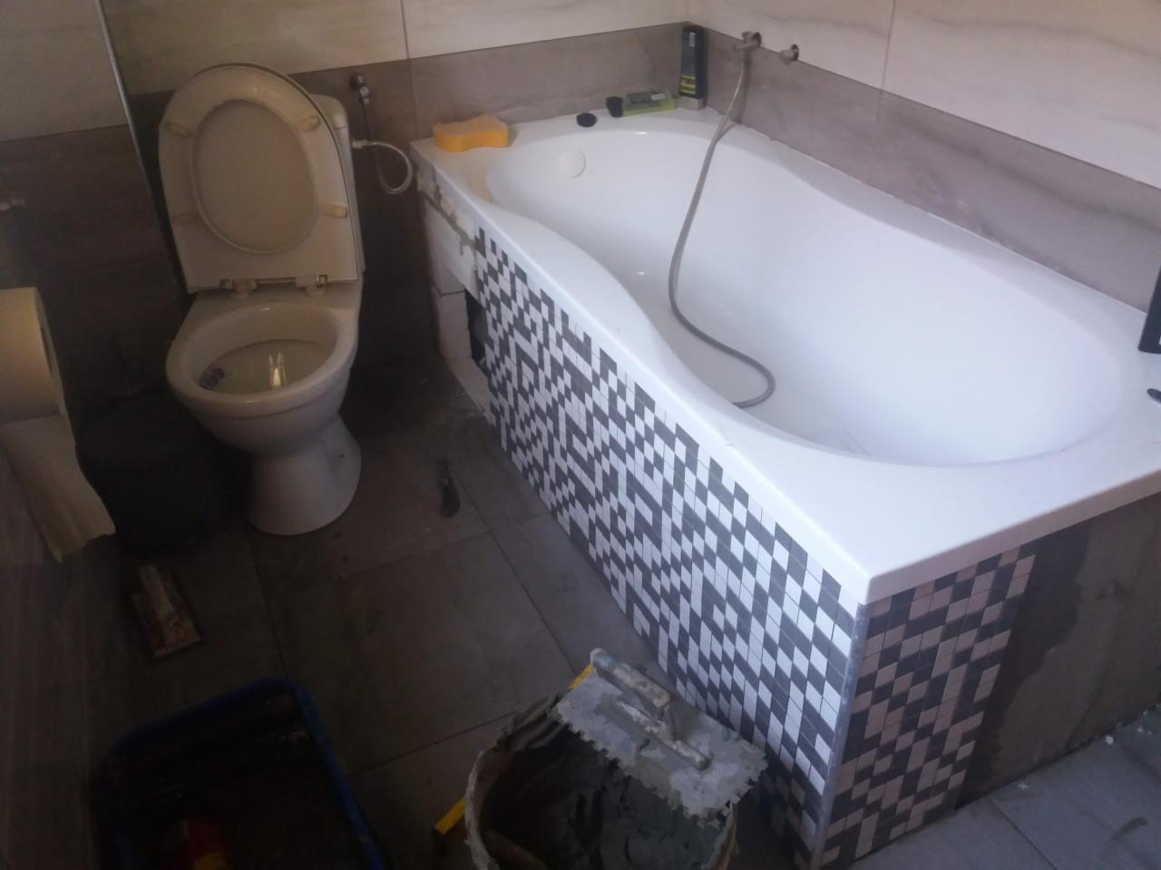 Biała wanna z mozaiką w czarno-białą szachownicę, toaleta z otwartą klapą, wiadro z zaprawą i kielnią na podłodze w trakcie prac wykończeniowych w łazience.