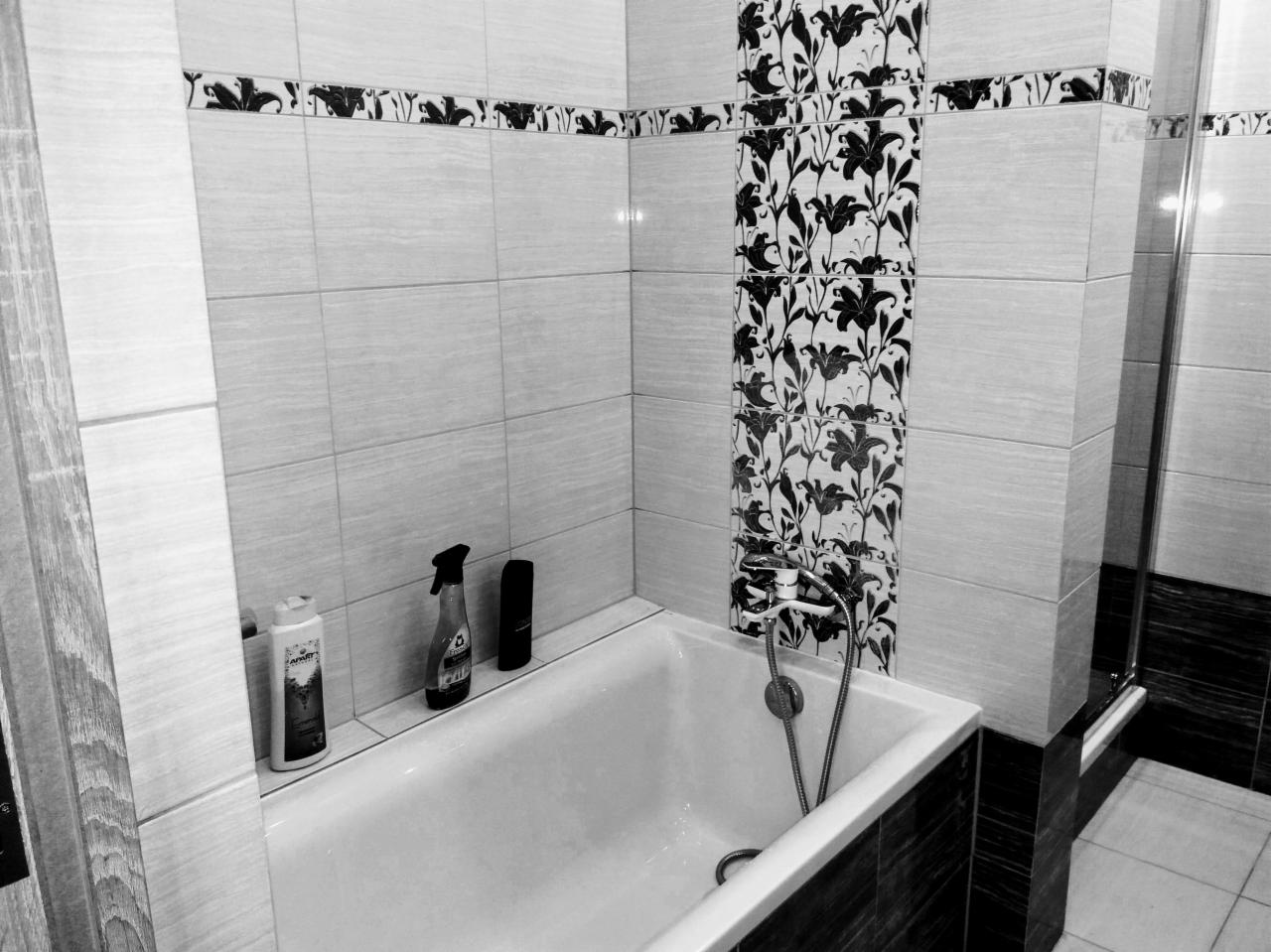 Wykończona łazienka w bieli i czerni z wanną, armaturą i dekoracyjnymi płytkami w kwiaty, widoczne butelki kosmetyków na rancie wanny.