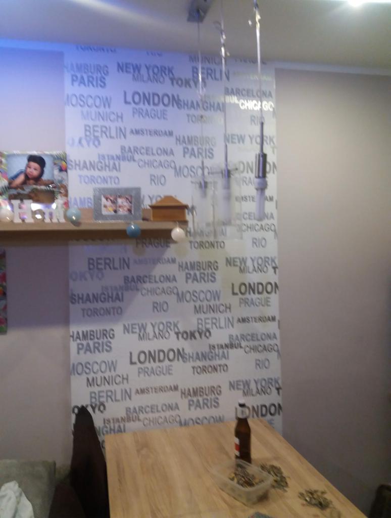 Ściana pokryta tapetą z nazwami miast, półka z ramkami i dekoracjami, fragment drewnianego stołu z butelką i drobnymi przedmiotami w pojemniku, oświetlenie sufitowe.