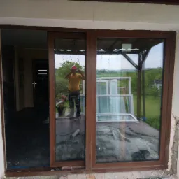 Brązowe okno przesuwne PCV w trakcie montażu, widoczne uszkodzenia ściany i odbicie osoby robiącej zdjęcie, na zewnątrz widoczne otoczenie domu i dodatkowe okna.