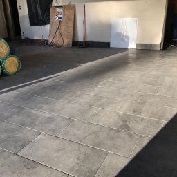 WALASEK-BUD - Ułożona szara, prostokątna podłoga z płytek w jasnym pomieszczeniu; w tle widoczne materiały izolacyjne i częściowo postać wchodząca do pomieszczenia.