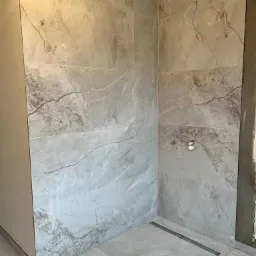 Wykończona kabina prysznicowa z szarych, marmurkowych płytek ceramicznych, widoczny odpływ liniowy i przygotowane przyłącza wodne.