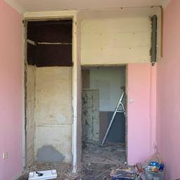 WALASEK-BUD - Pokój w trakcie demontażu: różowe ściany, zdemontowana szafa wnękowa z widoczną konstrukcją, gruz na podłodze i drabina oparta o ścianę w przejściu do kolejnego pomieszczenia.