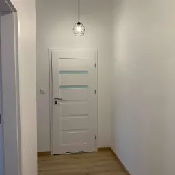 Wnętrze korytarza z drewnopodobną podłogą, białą ścianą i drzwiami wewnętrznymi z przeszkleniami, oświetlone wiszącą lampą w geometrycznej oprawie.