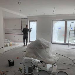 WALASEK-BUD - Malowanie natryskowe ścian w nowym wnętrzu; widoczne okna zabezpieczone folią, odsłonięte przewody oświetleniowe na suficie oraz sprzęt malarski.