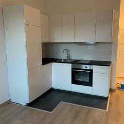 WALASEK-BUD - Nowoczesna, minimalistyczna kuchnia z białymi szafkami, czarnym blatem i ciemną podłogą, widoczna puszka z farbą na podłodze w rogu pomieszczenia.