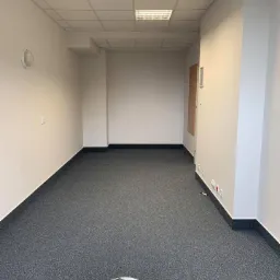 Wnętrze biura z nową szarą wykładziną dywanową, białe ściany z listwami przypodłogowymi w kolorze antracytowym, widoczny pojemnik z narzędziami do montażu wykładziny.