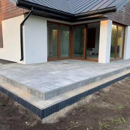 WALASEK-BUD - Nowoczesny taras z szarej kostki brukowej, z trzema stopniami i czarnym obrzeżem, przylegający do budynku z drewnianą elewacją i dużymi przesuwnymi oknami.