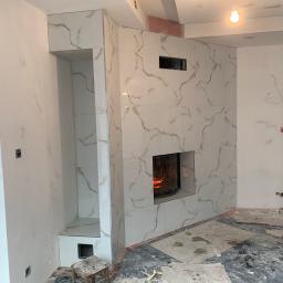 WALASEK-BUD - Kominek obłożony białymi płytkami imitującymi marmur, z paleniskiem i wnęką na drewno, w trakcie prac wykończeniowych, widoczne ślady po gładzi na ścianach i podłodze zabezpieczonej folią.