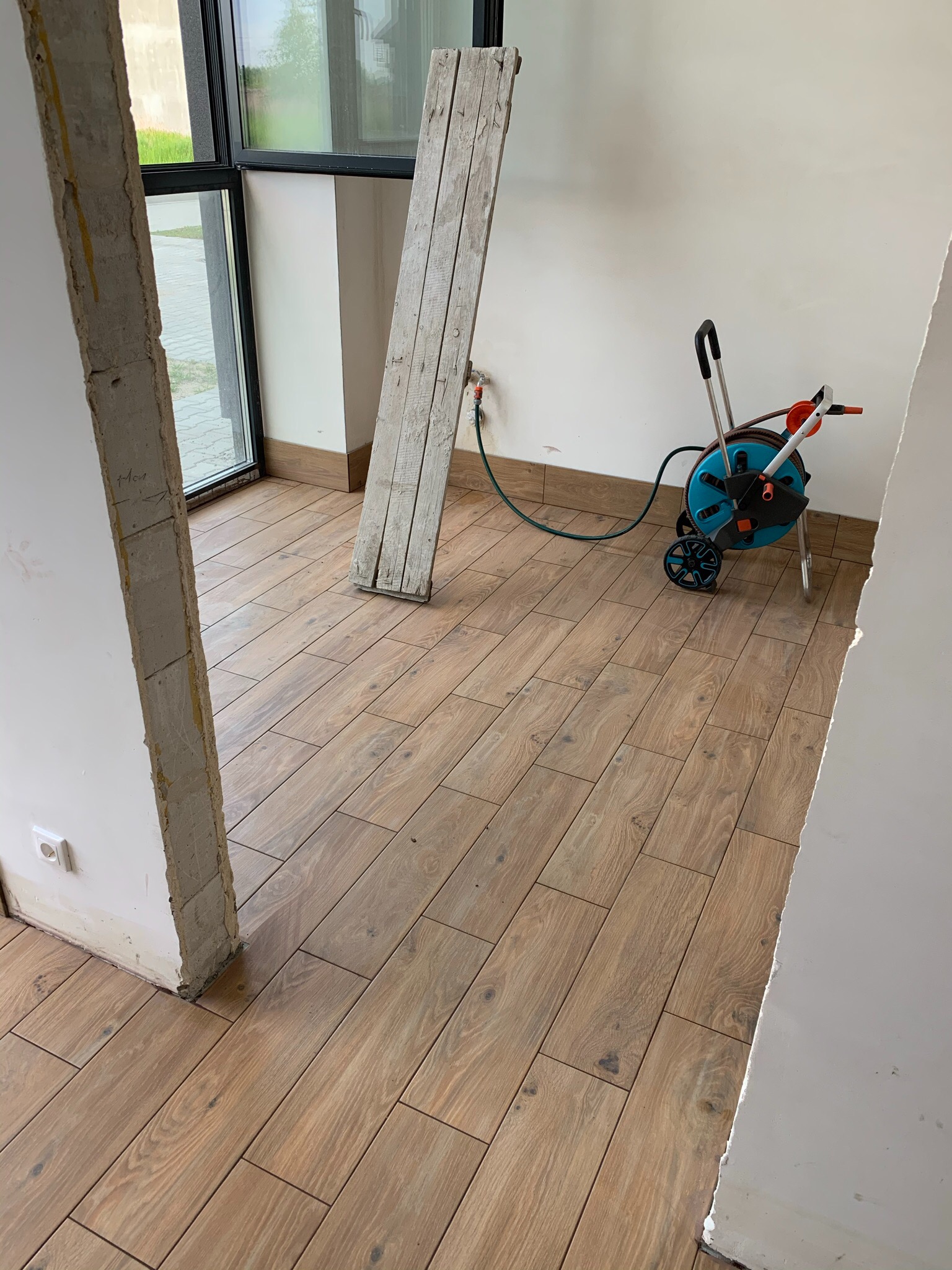 Ułożona drewnopodobna podłoga z płytek, widoczna szpara w ścianie, drabina oparta o ścianę i zwinięty wózek z wężem ogrodowym w rogu pomieszczenia.