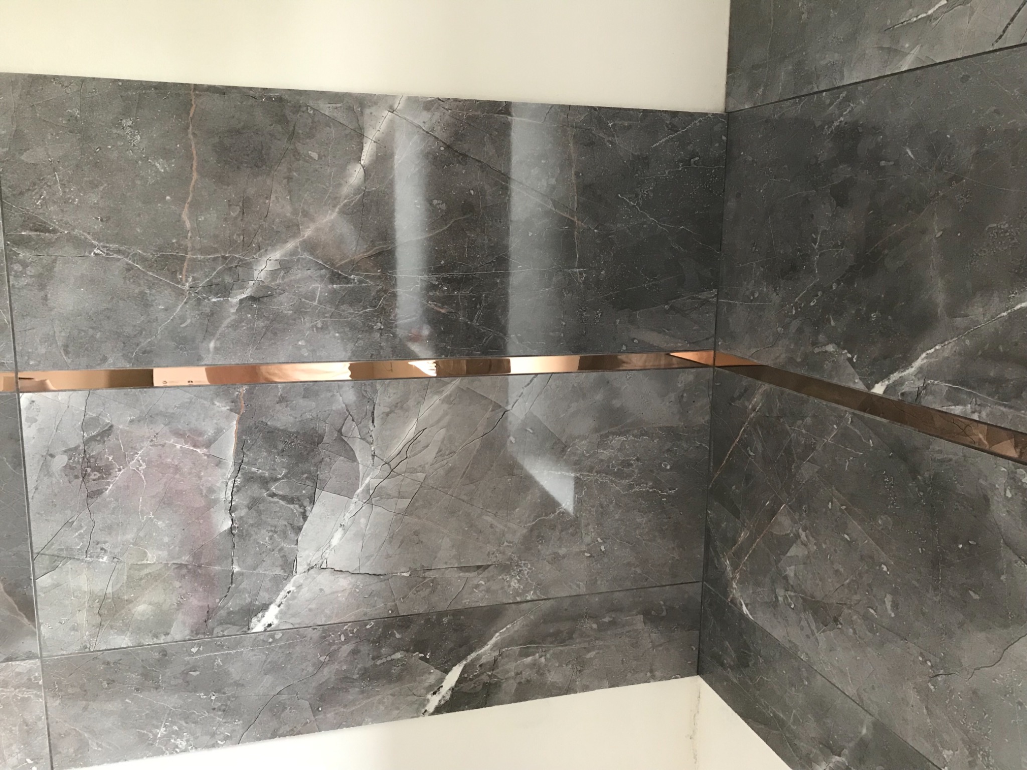 Fragment wykończonej ściany z szarych płytek imitujących marmur, z poziomym, miedzianym paskiem dekoracyjnym na wysokości wzroku.