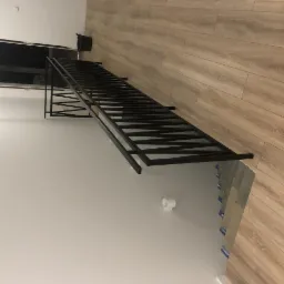 Częściowo zamontowana czarna metalowa balustrada schodowa w jasnym wnętrzu, widoczne elementy montażowe i surowe wykończenie podłogi.