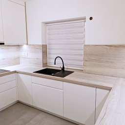 WITEK FURNITURE - Nowoczesna kuchnia z białymi szafkami, blatem imitującym drewno i czarnym zlewem z baterią, widoczne gniazdka elektryczne na ścianie.