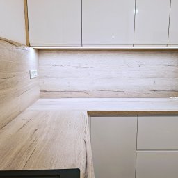 WITEK FURNITURE - Nowoczesna kuchnia z białymi, lakierowanymi szafkami górnymi i dolnymi, drewnianym blatem i panelem ściennym z widocznym włącznikiem światła i oświetleniem LED podszafkowym.