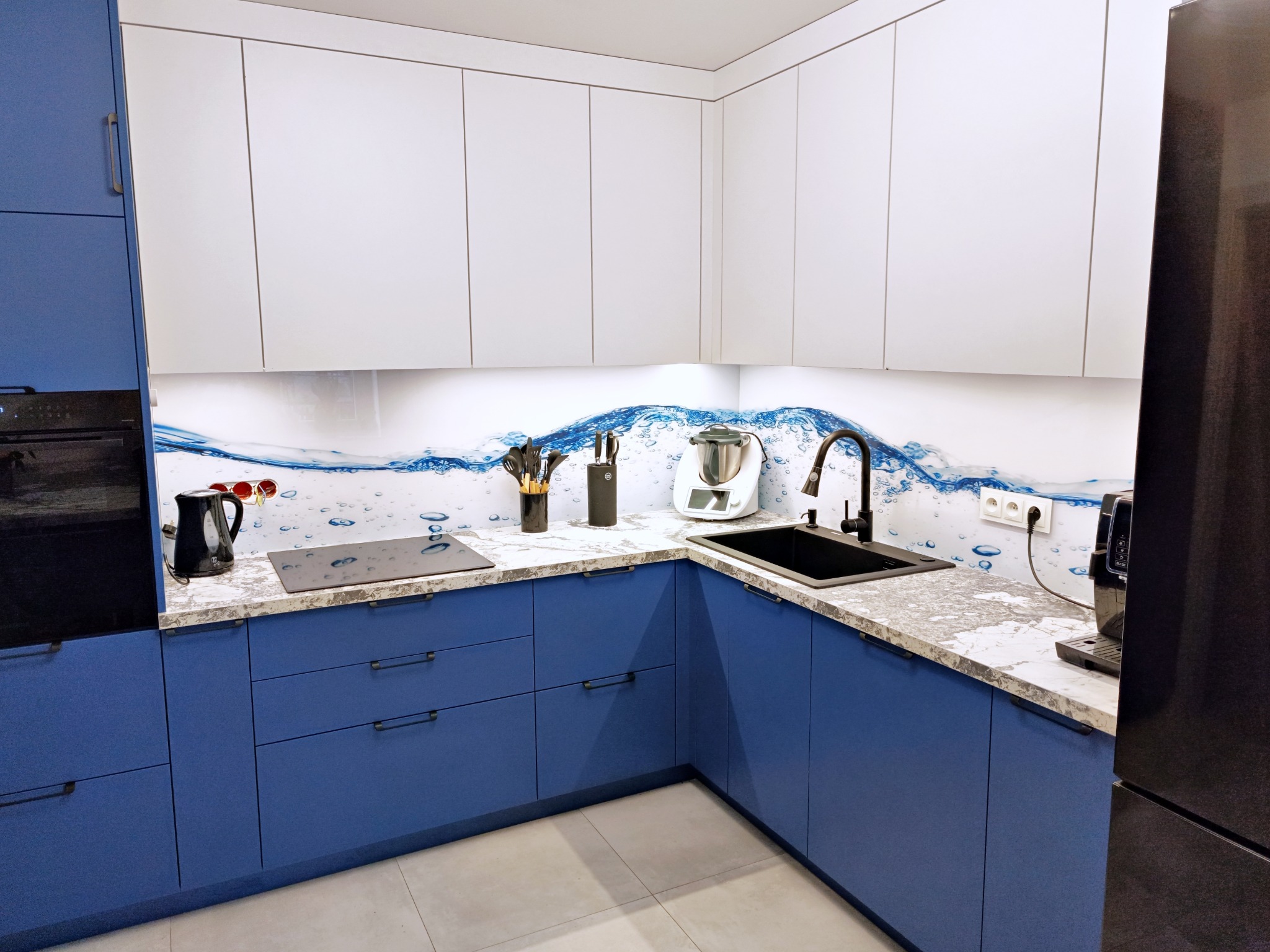 Nowoczesna kuchnia w odcieniach bieli i błękitu z blatem imitującym marmur, akcentowana grafiką z motywem wody na ścianie nad blatem, wyposażona w czarną armaturę i sprzęty AGD.