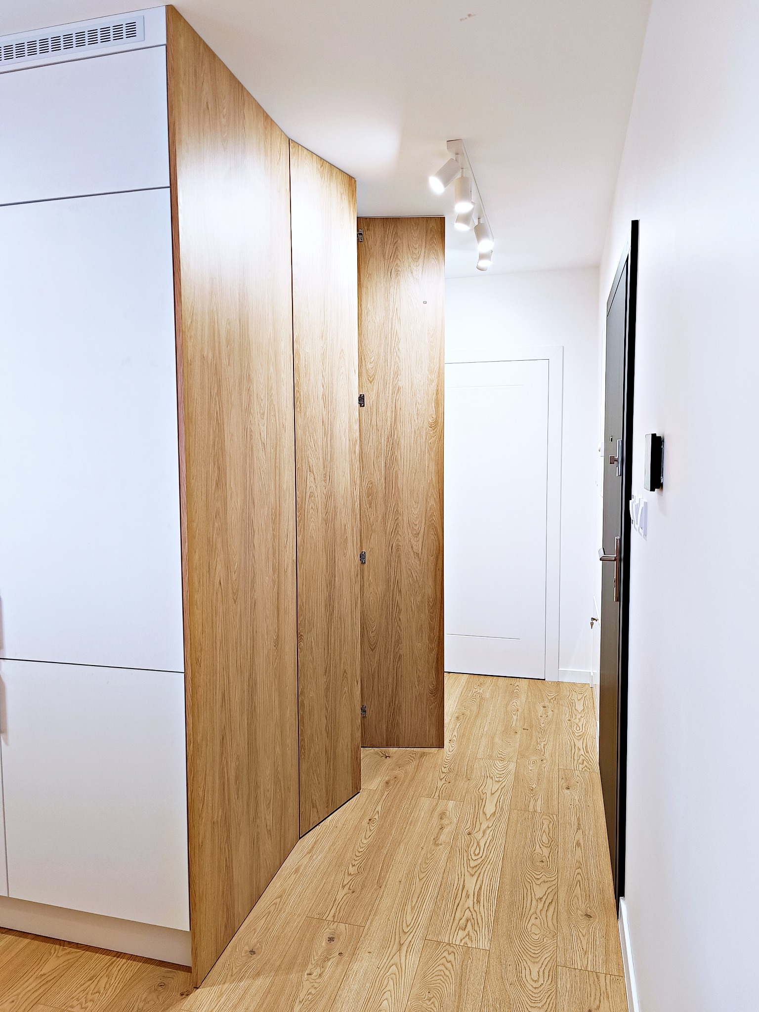 Wnętrze przedpokoju z zabudową: białe szafki i szafa z drewnianymi, fornirowanymi frontami, podłoga laminowana imitująca drewno, oświetlenie punktowe na suficie.