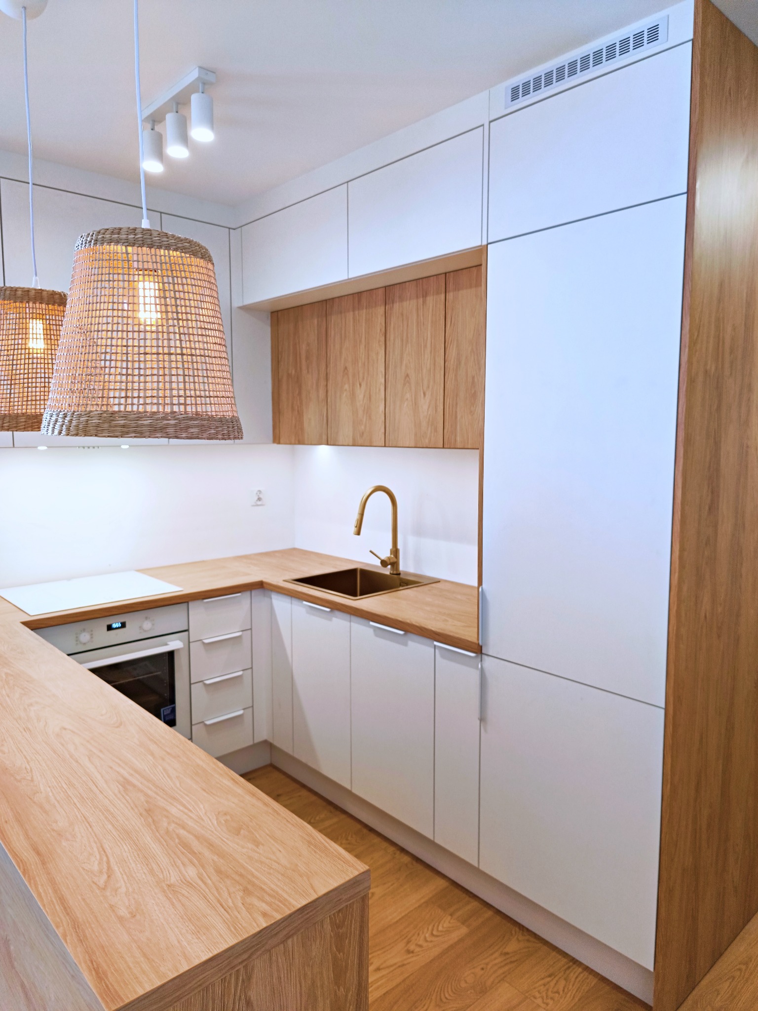 Nowoczesna kuchnia z białymi szafkami i drewnianymi blatami, złotą armaturą i plecionymi lampami wiszącymi