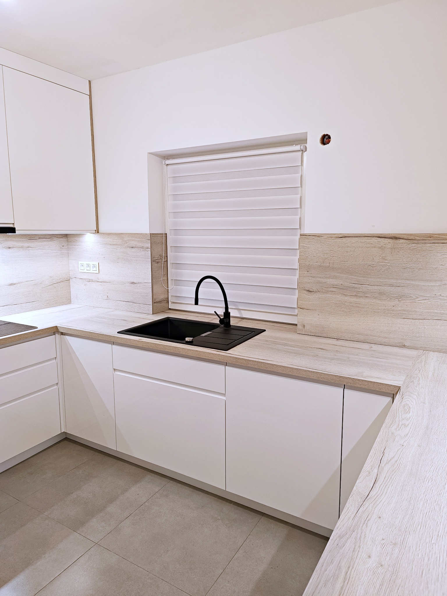 Nowoczesna kuchnia z białymi szafkami, blatem imitującym drewno i czarnym zlewem z baterią, widoczne gniazdka elektryczne na ścianie.