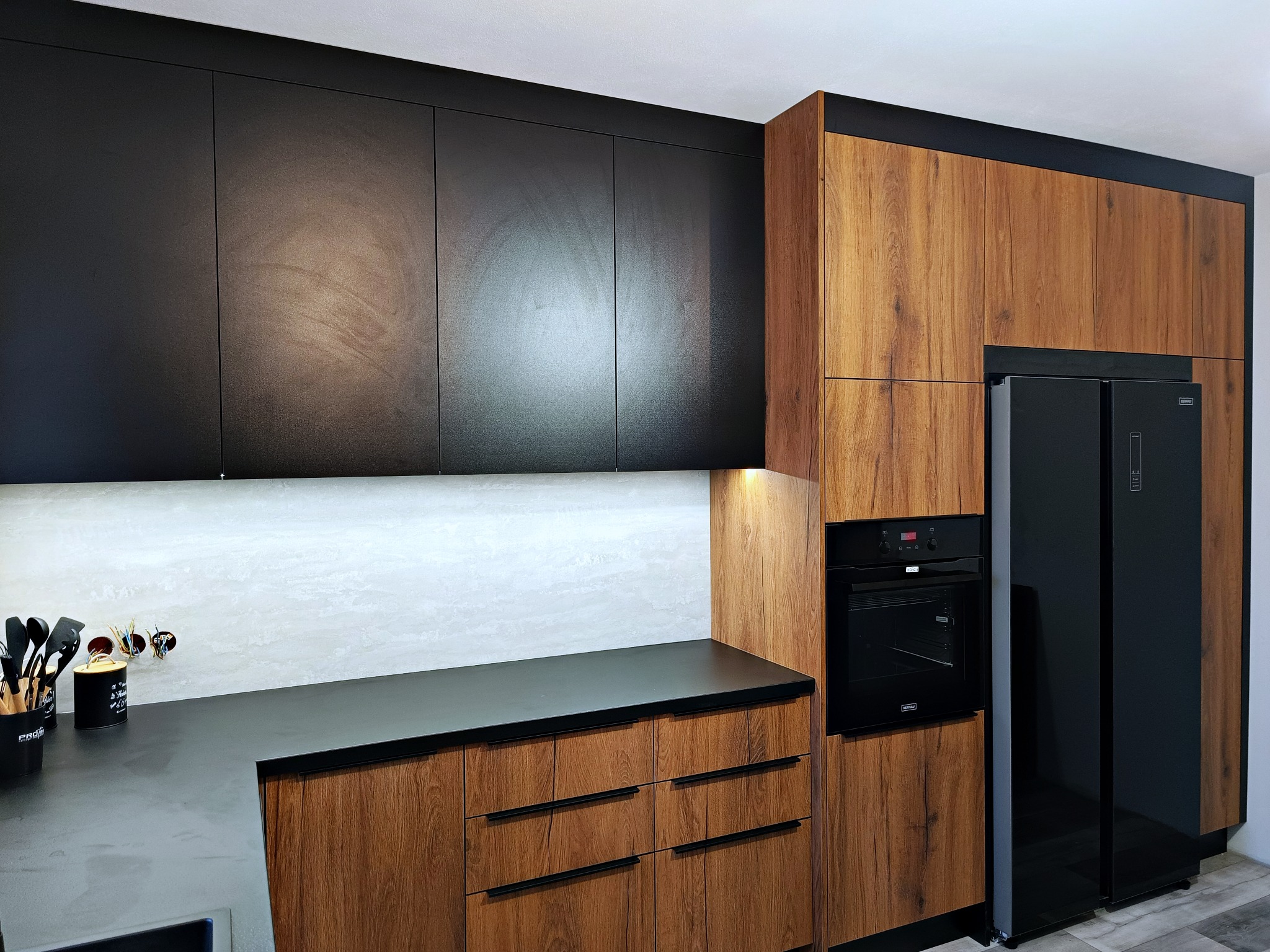 Nowoczesna kuchnia z czarnymi górnymi szafkami i drewnopodobnymi dolnymi, zintegrowana z czarnym piekarnikiem i lodówką, widoczne akcesoria kuchenne na blacie.