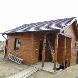 Dach Pol Zieliński - Drewniany domek z nowym dachem pokrytym ciemną dachówką, widoczna drabina oparta o ścianę i elementy wykończeniowe obok wejścia.