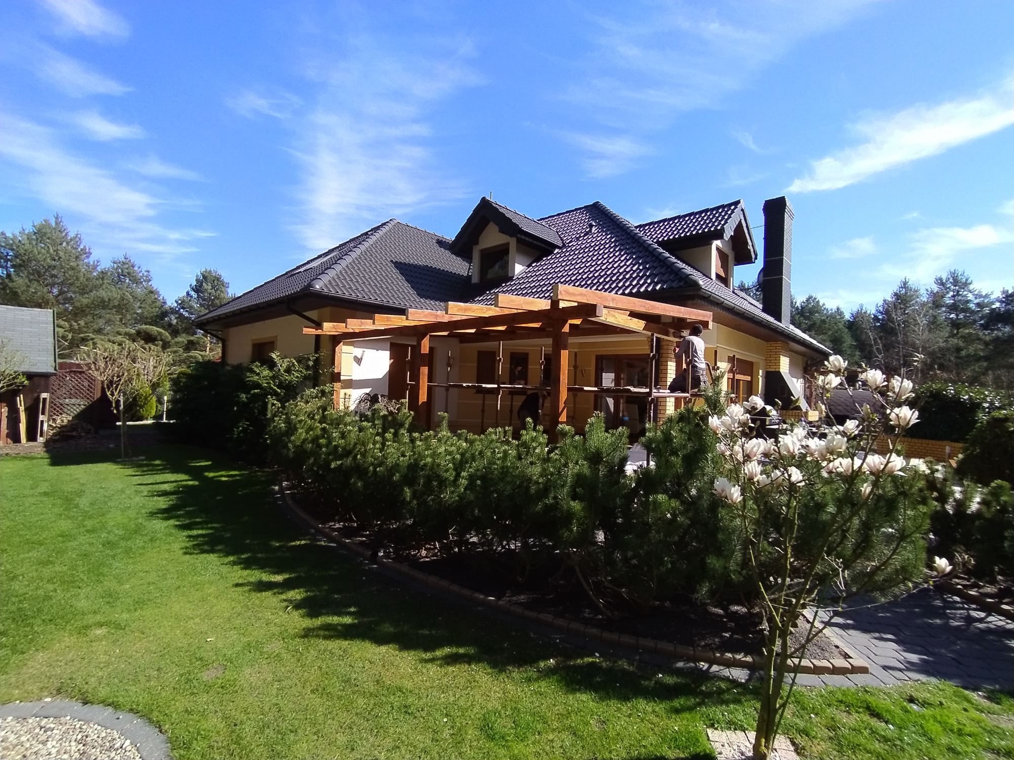 Dom jednorodzinny z ciemną dachówką i drewnianą pergolą w trakcie montażu, widoczne rusztowania i pracownicy, w tle błękitne niebo z delikatnymi chmurami.