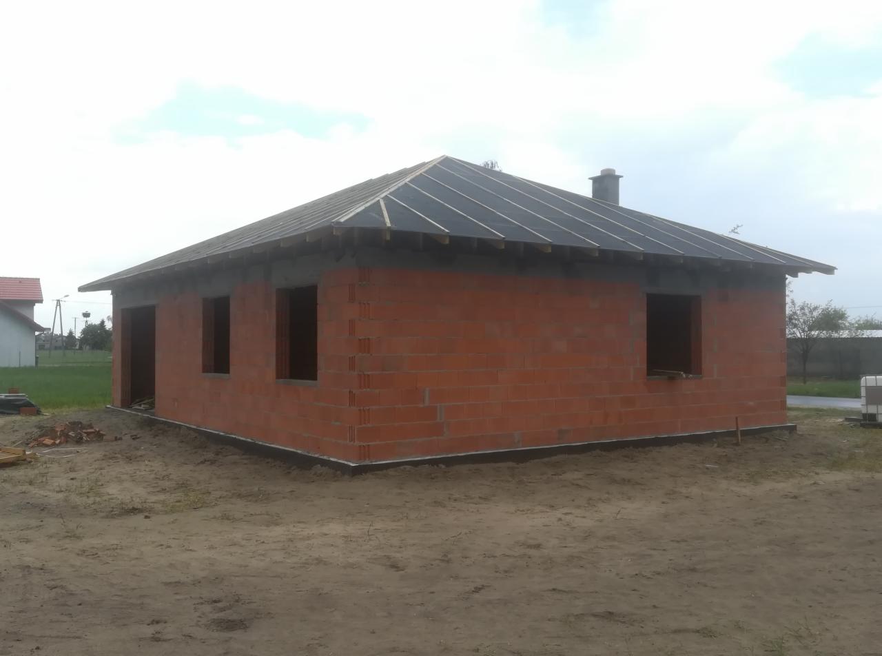 Niedokończony budynek z czerwonej cegły z przygotowaną konstrukcją pod pokrycie dachu, widoczne łaty dachowe i komin, otoczenie piaszczyste.