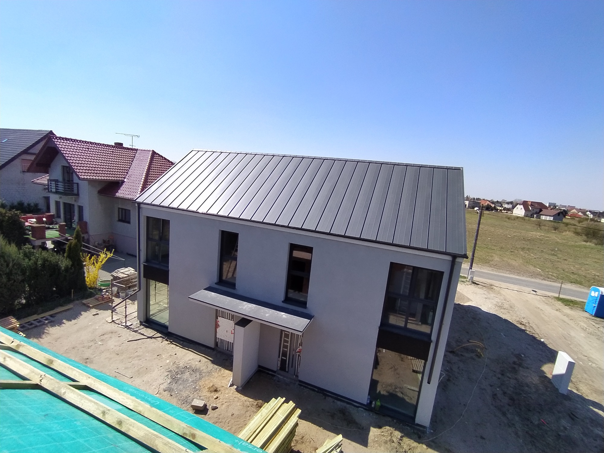 Nowoczesny dom jednorodzinny z grafitowym dachem z blachy trapezowej, widok z góry na plac budowy z elementami więźby dachowej w pierwszym planie.