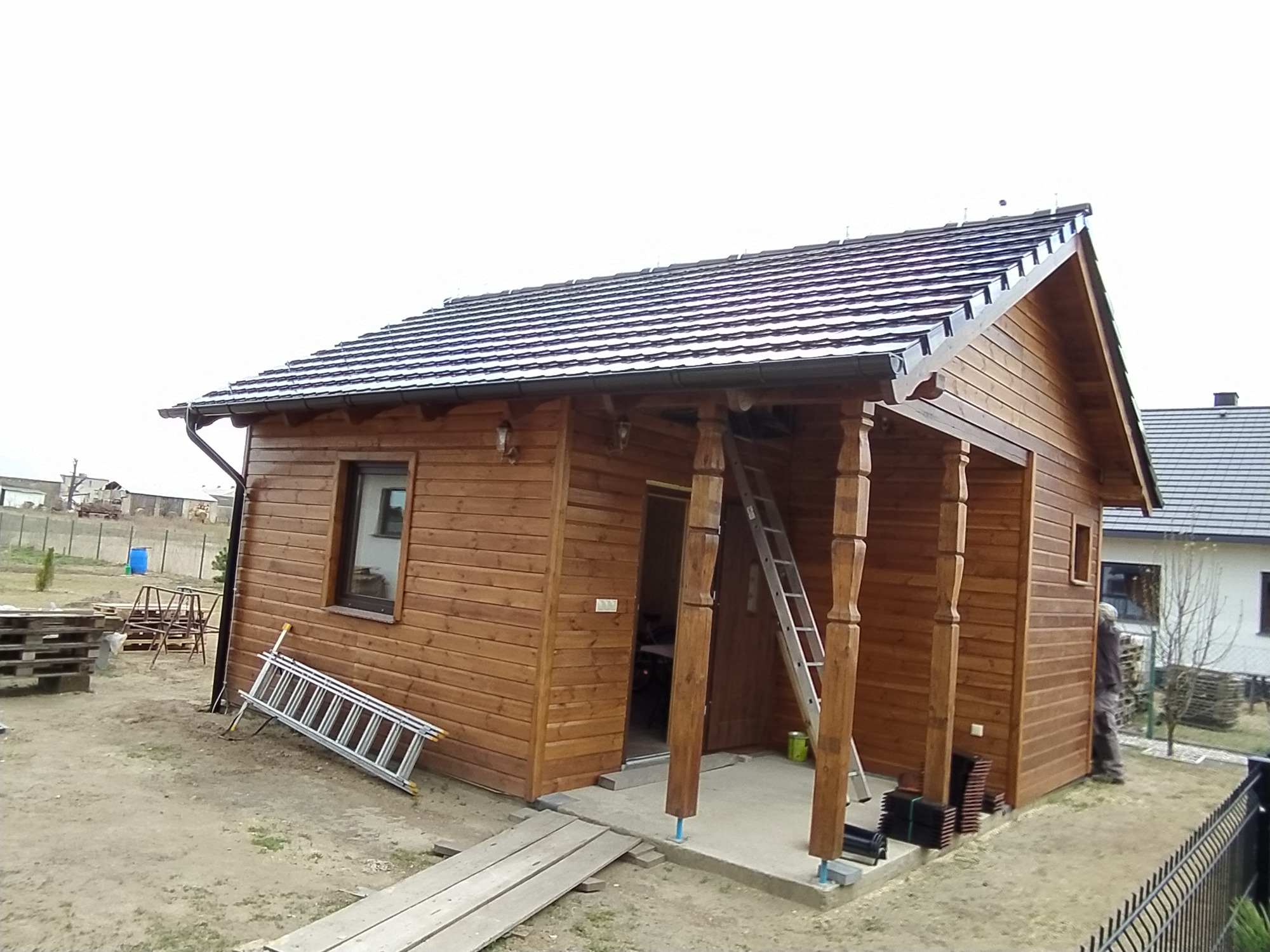 Drewniany domek z nowym dachem pokrytym ciemną dachówką, widoczna drabina oparta o ścianę i elementy wykończeniowe obok wejścia.
