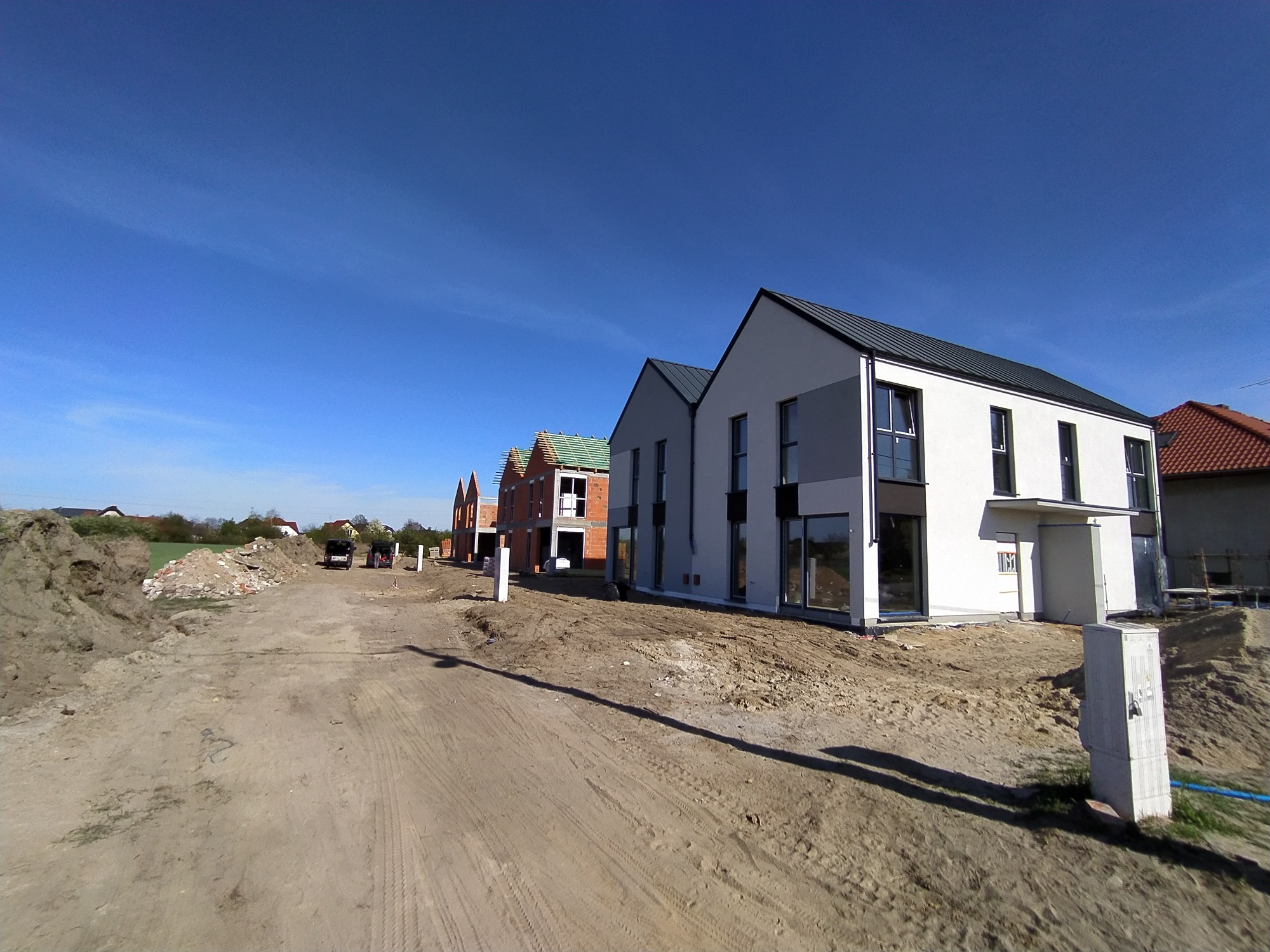 Nowe domy jednorodzinne w Witkowie w trakcie budowy, widoczne z perspektywy drogi gruntowej, z różnymi etapami prac na poszczególnych budynkach, od stanu surowego do wykończonego z ciemną blachą...