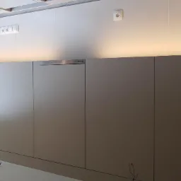 Szare, minimalistyczne szafki kuchenne z ukrytymi uchwytami i podświetleniem LED, z widoczną otwartą półką regału po lewej stronie i wystającymi kablami na podłodze.