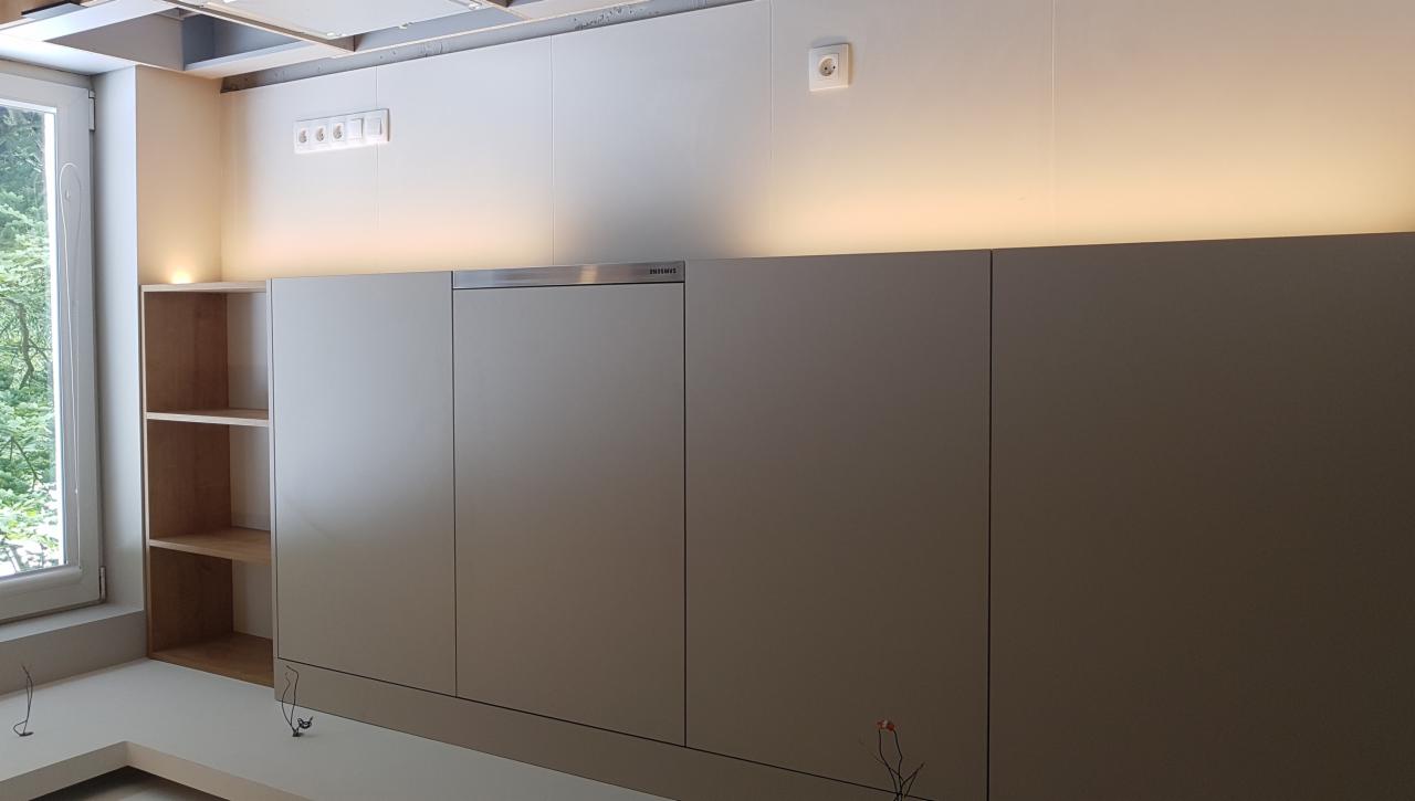 Szare, minimalistyczne szafki kuchenne z ukrytymi uchwytami i podświetleniem LED, z widoczną otwartą półką regału po lewej stronie i wystającymi kablami na podłodze.