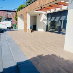 Nowoczesna elewacja domu z jasnym tynkiem, drewnianymi elementami dekoracyjnymi i tarasem wyłożonym płytkami imitującymi drewno, schody wejściowe z szarego kamienia.