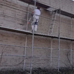 Pracownik na rusztowaniu aplikuje tynk maszynowy na ścianę budynku z cegły, widoczne narzędzia i niezagospodarowany teren u podstawy ściany.