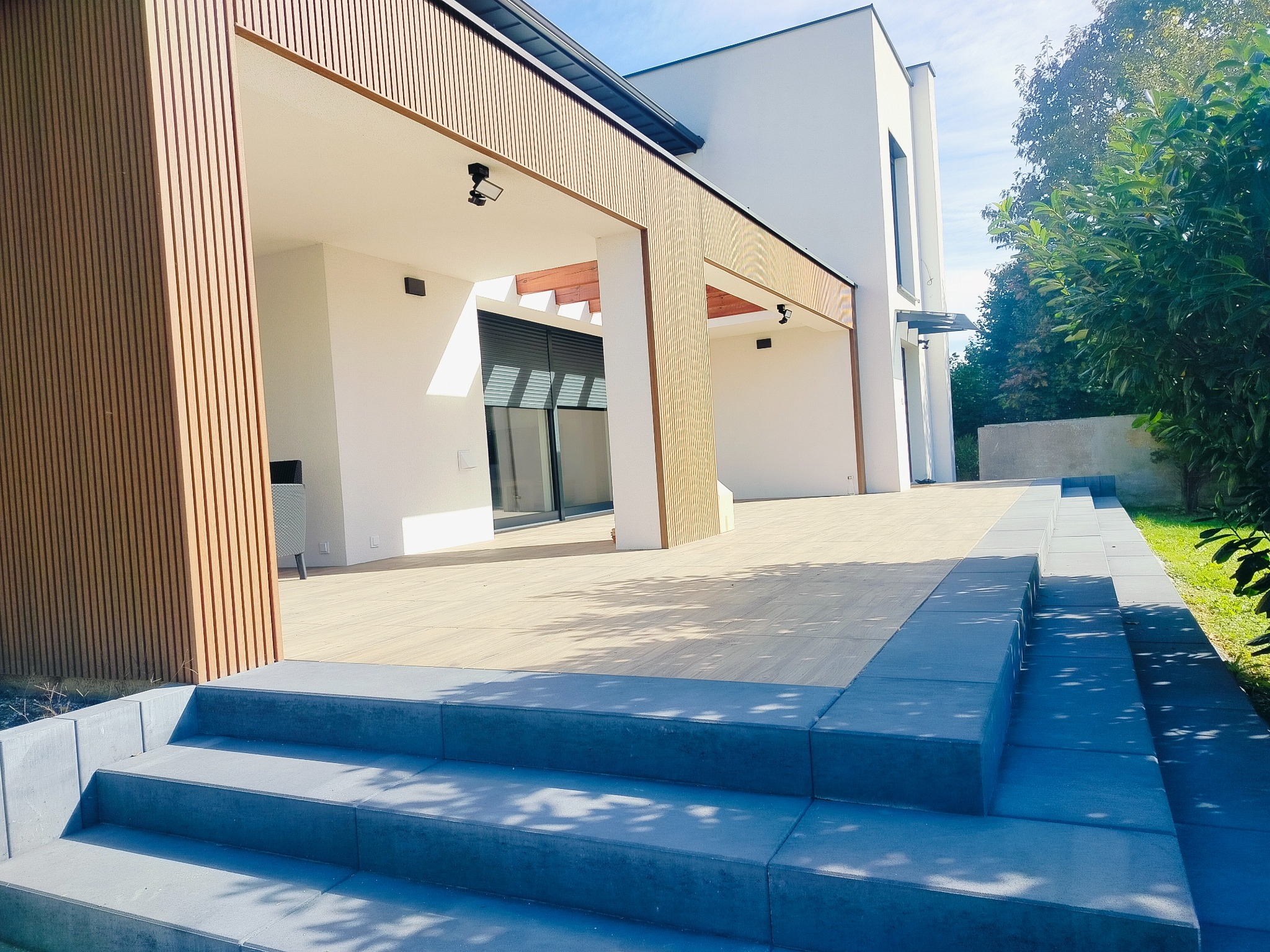 Nowoczesny taras z drewnianą elewacją i schodami z szarej kostki brukowej w słoneczny dzień.