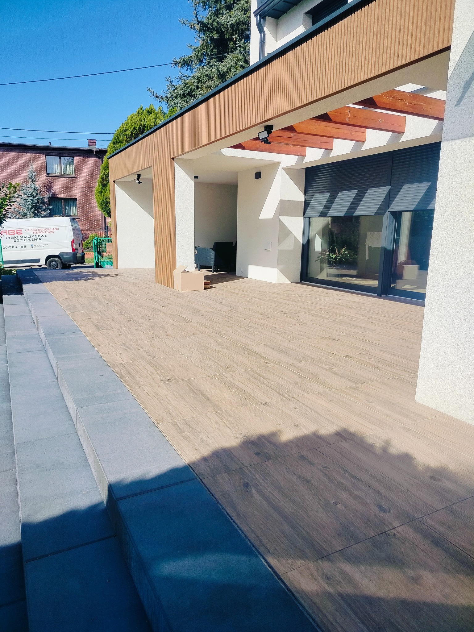 Nowoczesna elewacja domu z jasnym tynkiem, drewnianymi elementami dekoracyjnymi i tarasem wyłożonym płytkami imitującymi drewno, schody wejściowe z szarego kamienia.