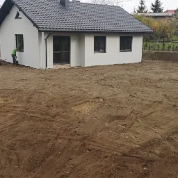 Nowo wybudowany, otynkowany dom jednorodzinny z ciemną dachówką, otoczony przygotowanym terenem pod ogród, gdzie pracownik z łopatą wyrównuje ziemię przy fundamencie. Widoczne ślady opon po maszynach.