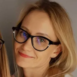 Portret uśmiechniętej blondynki w okularach, z widocznym kolczykiem w uchu i drugim zestawem okularów w tle.